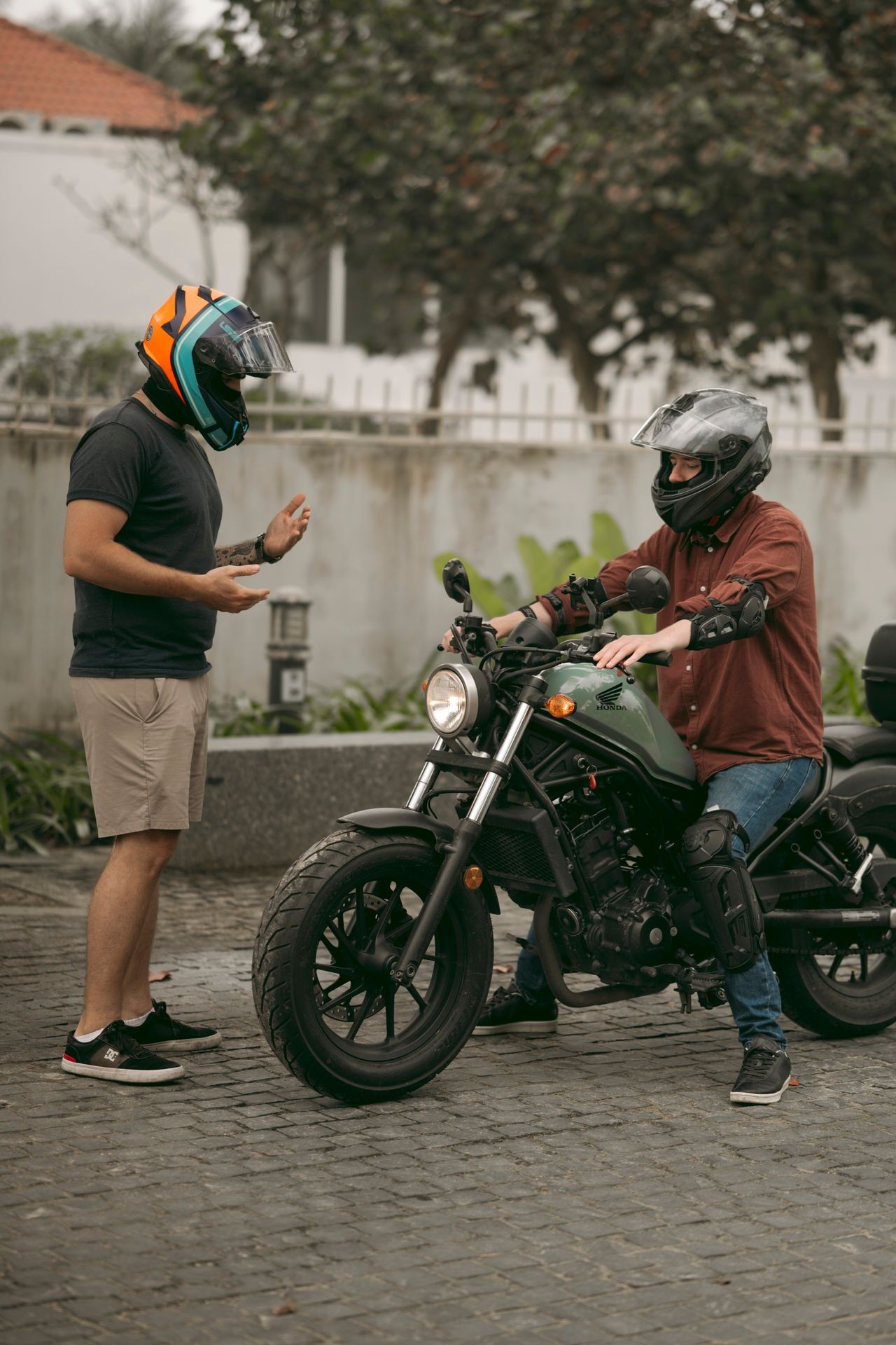 Two people wearing helmets, one standing and one sitting on a motorcycle, having a conversation.