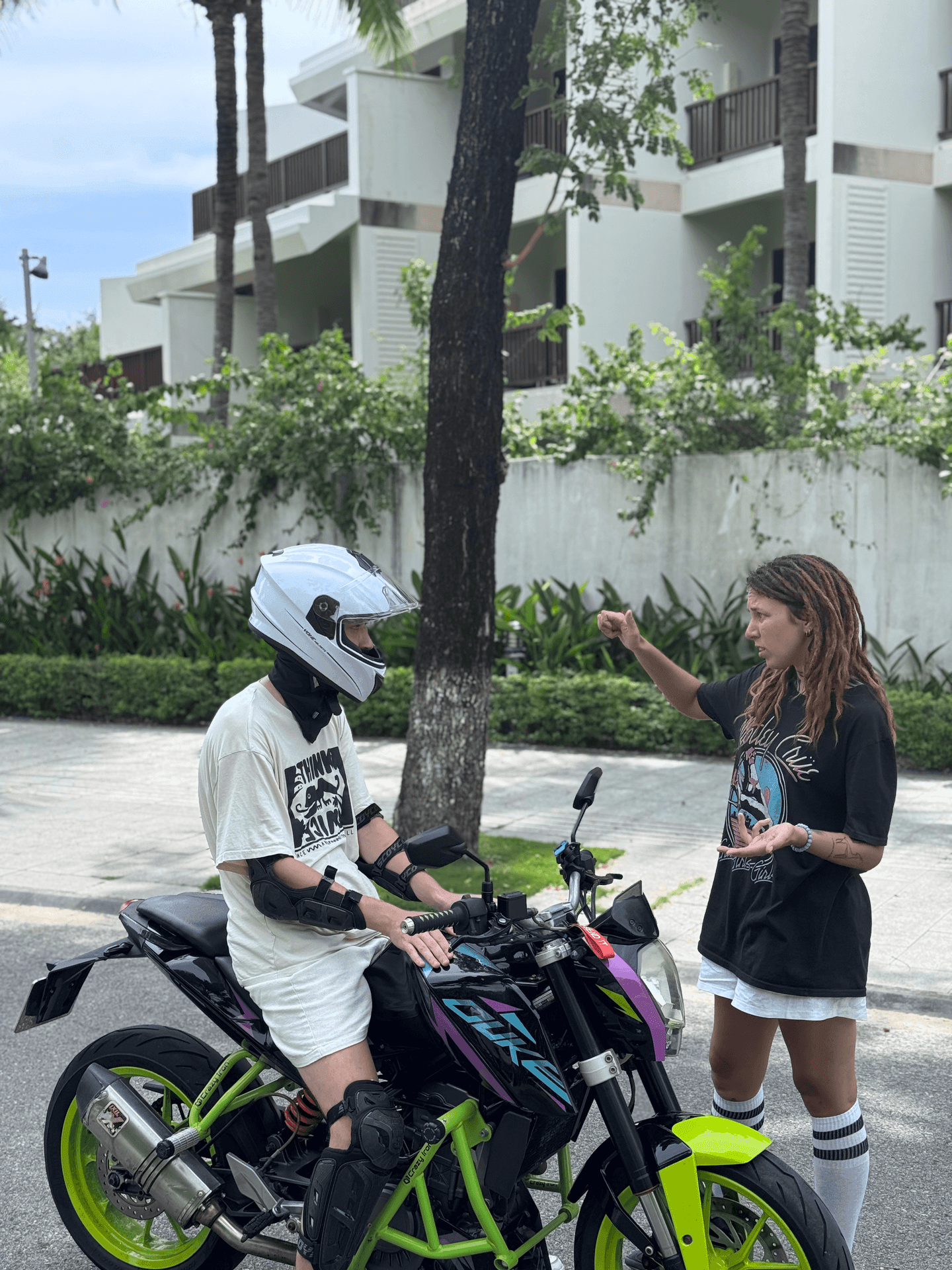 Person on motorcycle with helmet talking to another person on a sidewalk, residential building in background.