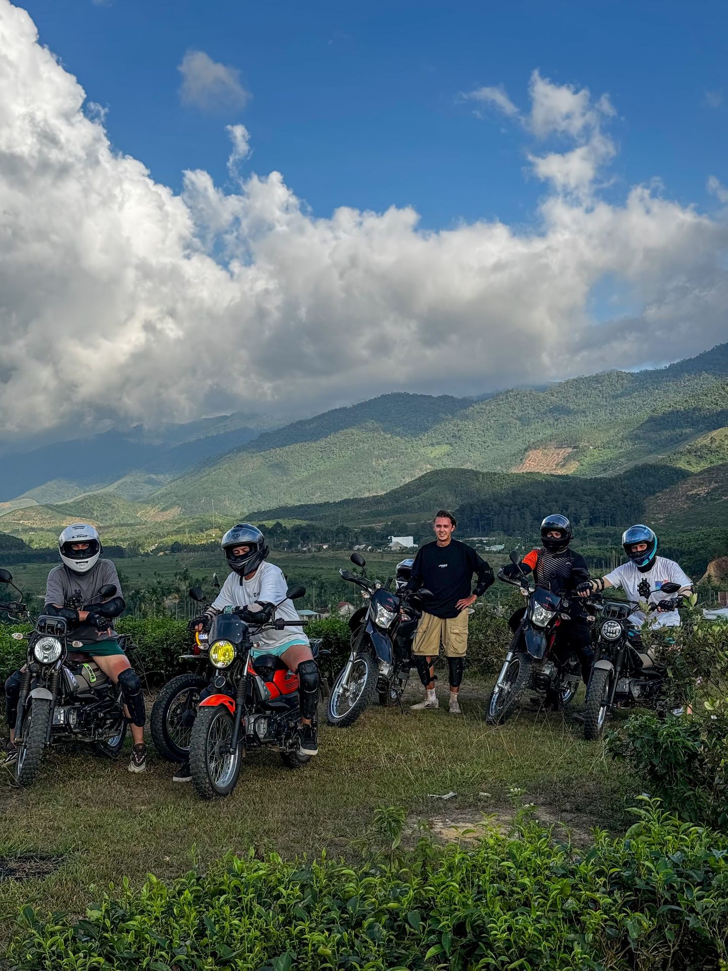 Group of people with dirt bikes posing on a grassy hill with a backdrop of mountains and clouds.